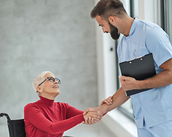 An old woman in a wheelchair holds the arm of a male nurse holding a binder